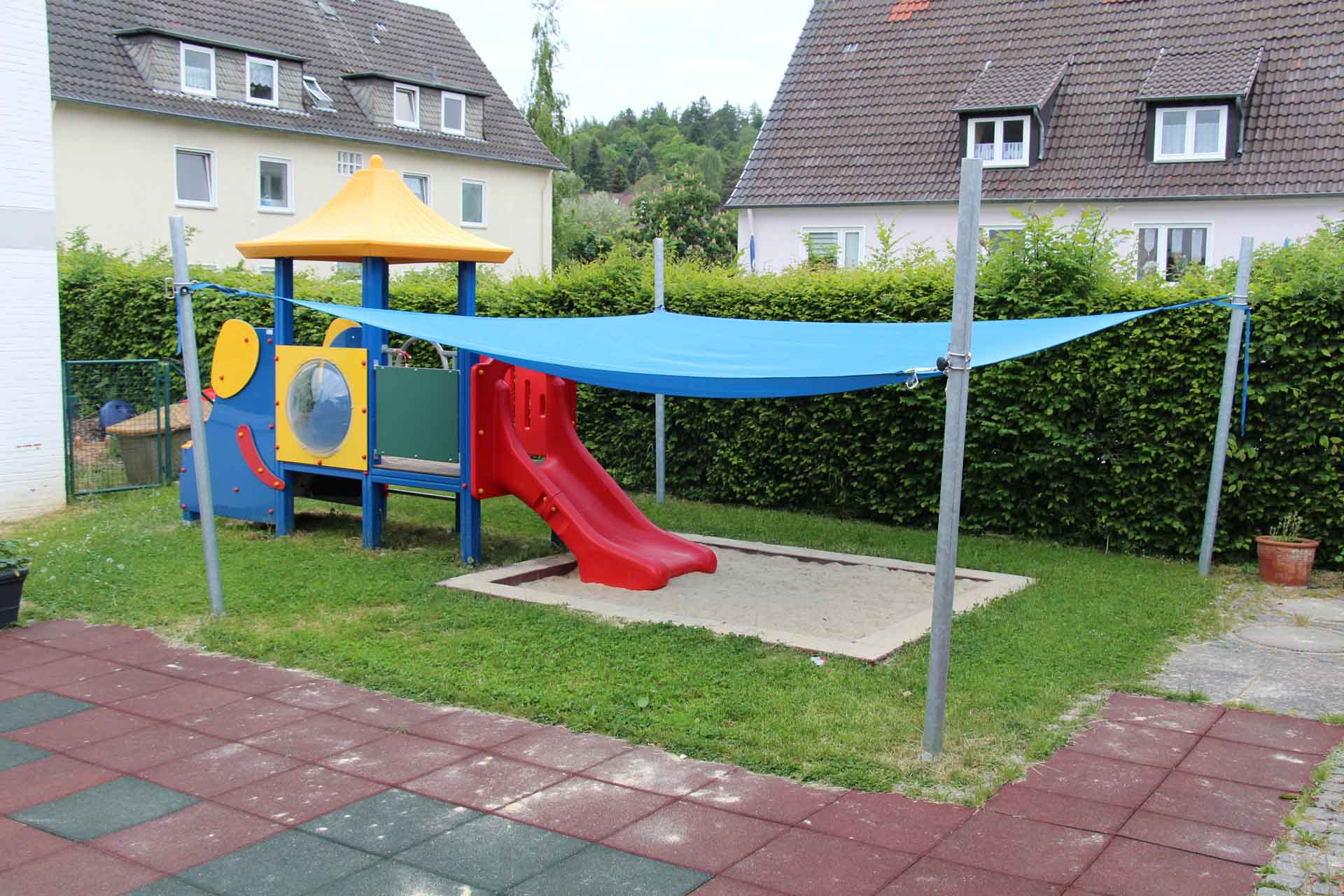 Kleiner Spielplatz mit Rutsche, Sandkasten und blauem Sonnensegel.